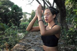 A woman in a forest practicing qigong healing with hands in prayer position over the forehead
